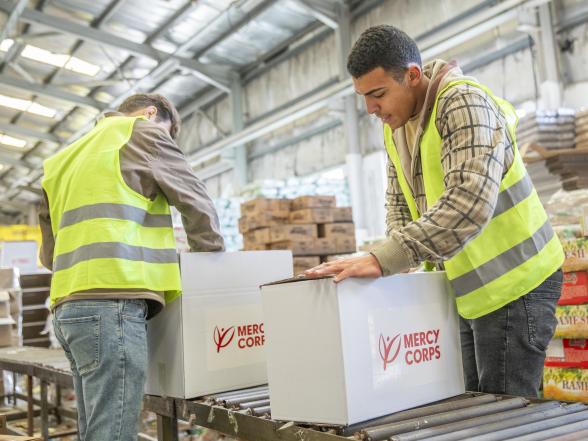 Two team members wearing safety vests package goods in boxes labeled with the mercy corps logo.