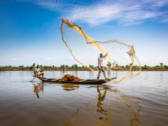 Fishermen in a small wooden boat cast a large yellow net against a blue sky