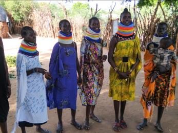 Six kenyan women, one of whom holding a toddler, stand together in their village.
